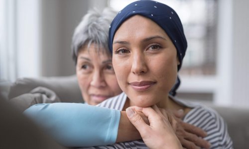 An ethnic woman wearing a headscarf and fighting cancer sits on the couch with her mother. She is in the foreground and her mom is behind her, with her arm wrapped around in an embrace. She is looking at the camera with an expression of resolute confidence and serenity.